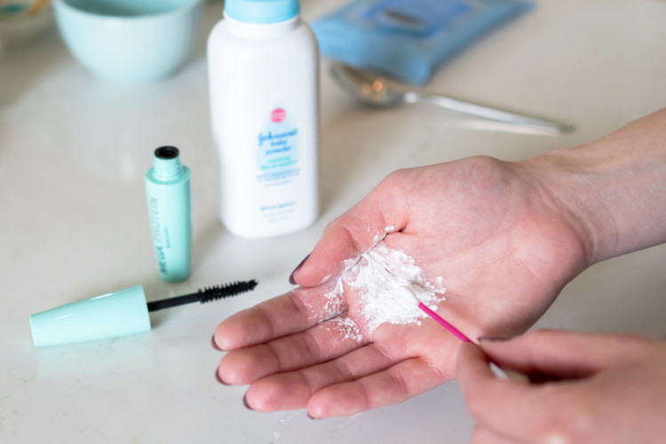 Baby powder in the palm of a persons hand being picked up on a Q-tip with a tube of mascara and the baby powder bottle on the counter in the background